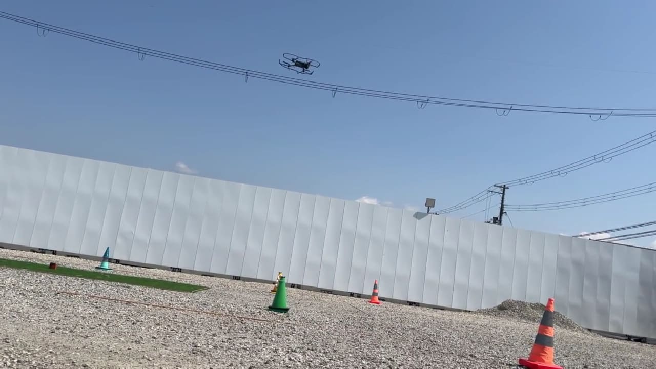 講習中、白いフェンスの上空を飛ぶドローン（動画のポスター画像）
